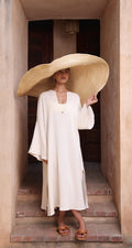 A woman stands in a doorway wearing Thr3letter's flowing Kiaan Kaftan in a soft, neutral almond color.